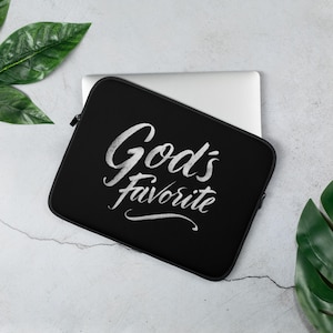Puede incluir: Funda negra para portátil con el texto "God's Favorite" en escritura blanca. La funda tiene cierre de cremallera. La funda para portátil está sobre una superficie gris claro con hojas verdes.