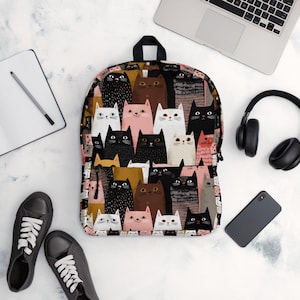 以下が含まれることがあります： 黒、白、ピンク、茶色など、さまざまな色の様式化された猫の繰り返し模様が特徴のバックパック。 バックパックには黒いハンドルとストラップが付いています。 背景はピンク、黒、金のミックスです。