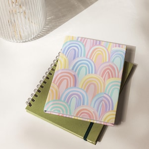 May include: Two notebooks. One spiral-bound notebook with a green cover and a second notebook with a cover featuring a pastel rainbow pattern. The notebooks are stacked on a white surface, with a glass vase in the background.