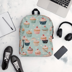 May include: A light blue backpack with a pattern of cupcakes with cherries, on a white polka dot background. The backpack has a black handle and zipper. Black sneakers, a laptop, headphones, a phone, and a notebook are also in the image.