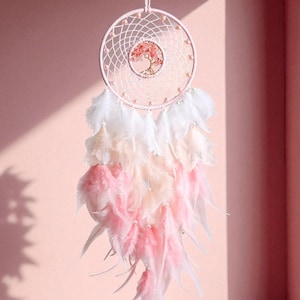 Puede incluir: Un atrapasueños rosa con un diseño de árbol en el centro. El atrapasueños presenta un marco circular con una red tejida y está adornado con plumas blancas, melocotón y rosas. Se adjunta una cinta rosa para colgarlo.