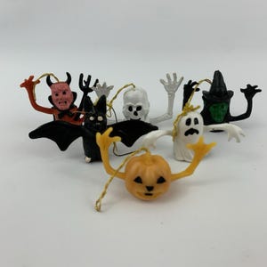 May include: A collection of six small Halloween-themed hanging ornaments featuring a red devil, a black bat, a white skeleton, a white ghost, a green-faced witch in a black hat, and an orange jack-o'-lantern, each with a gold hanging string.