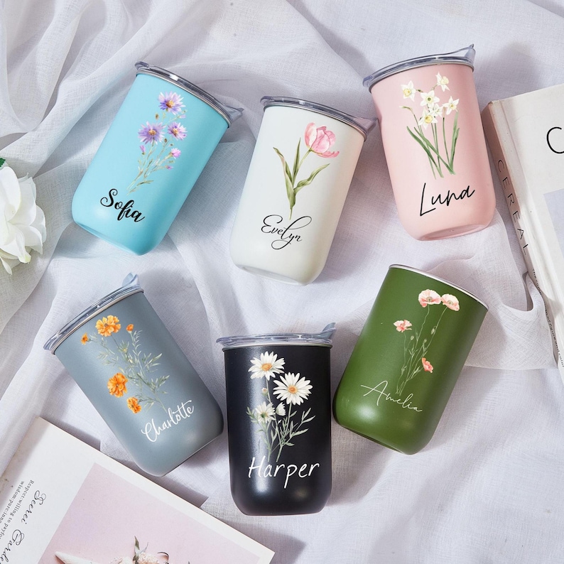 May include: A collection of personalized stainless steel tumblers in various colors, each adorned with floral designs and a name. Colors include light blue, white, pink, gray, black, and green. The names Sofia, Evelyn, Luna, Charlotte, Harper, and Amelia are printed on the tumblers.