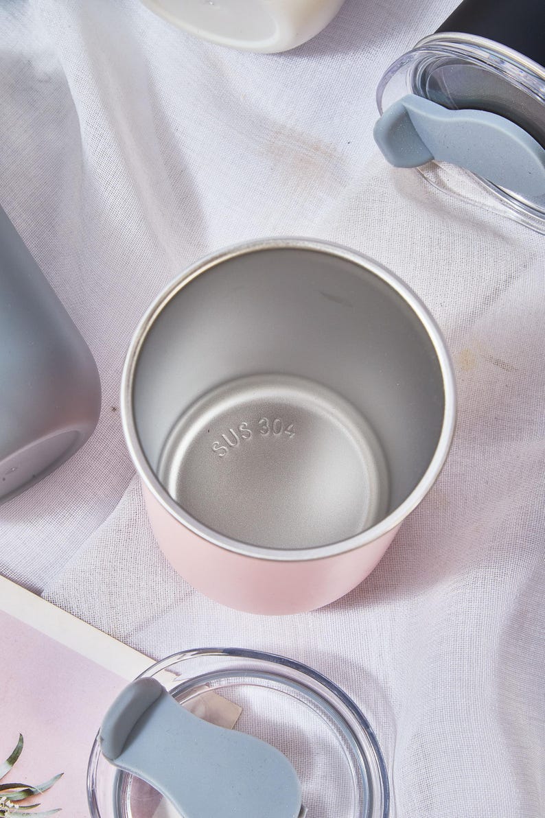 May include: A pink insulated tumbler with a silver interior and a clear lid with a gray silicone piece. The bottom of the tumbler is stamped with "SUS 304". Other tumblers and lids are visible.
