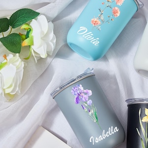 May include: A collection of personalized tumblers in various colors, each adorned with floral designs and names. The tumblers are light blue, gray, and black, with names like "Olivia" and "Isabella" in white script. The floral designs include orange and purple flowers.