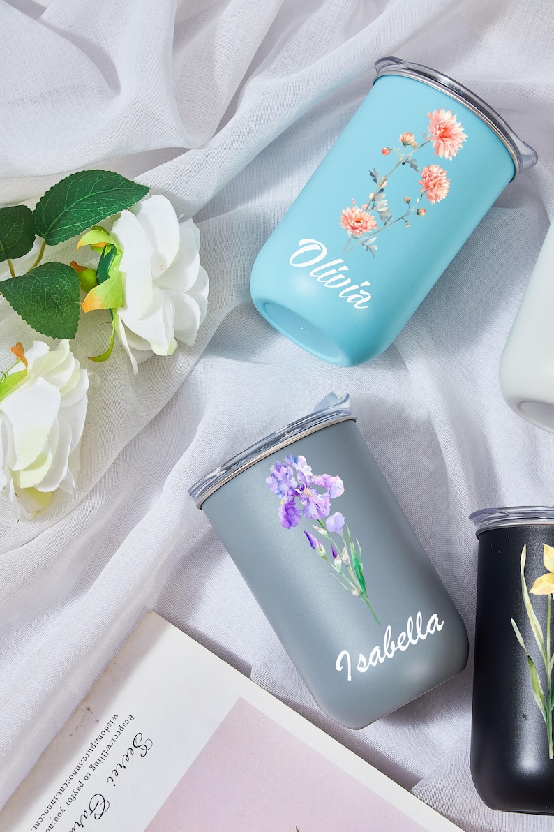 May include: A collection of personalized tumblers in various colors, each adorned with floral designs and names. The tumblers are light blue, gray, and black, with names like "Olivia" and "Isabella" in white script. The floral designs include orange and purple flowers.