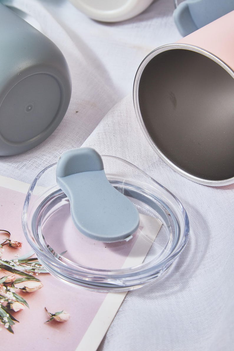 May include: Close-up of several insulated tumblers and lids. One tumbler is pink, another is gray, and a third is white. The clear lid has a light blue slider. The tumblers are on a white cloth.