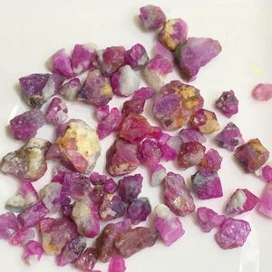 May include: A collection of raw, unpolished ruby gemstones in various shades of vibrant pink and deep magenta. These small, irregular mineral fragments feature translucent to opaque textures with occasional white and tan inclusions throughout.
