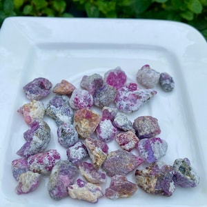 Peut inclure: Une collection de pierres précieuses brutes, non taillées, dans des tons de rose, de violet et de gris, disposées sur une assiette carrée blanche. Les pierres varient en taille et en forme, avec des structures cristallines visibles. Fond vert flou.