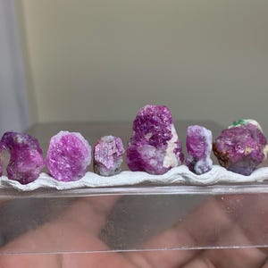 Peut inclure: Une collection de huit cristaux de pierres précieuses brutes, d'un magenta vibrant et d'un rose profond, disposés en rangée horizontale. Chaque pierre présente une texture naturelle rugueuse et est fixée sur une fine bande adhésive blanche.