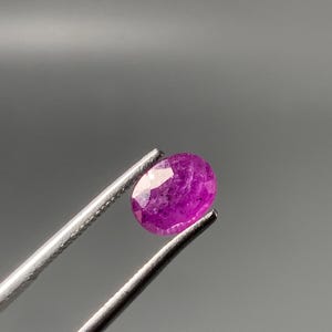Puede incluir: Una gema de color magenta con corte ovalado sostenida por unas pinzas de metal plateado. La piedra presenta una superficie facetada que refleja la luz, mostrando un tono rosa violáceo intenso y una estructura cristalina translúcida.