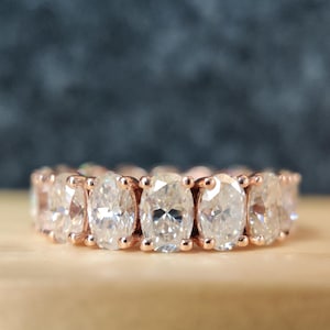 Pode incluir: Anel de eternidade em ouro rosa com uma série de pedras preciosas de corte oval. As pedras são colocadas próximas umas das outras, criando uma faixa contínua de brilho. O anel é exibido em uma superfície de madeira clara com um fundo escuro.
