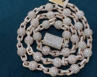 May include: A rose gold chain necklace with a series of round, diamond-encrusted beads connected by smaller links. The necklace features a rectangular clasp also covered in diamonds. The jewelry is displayed on a dark teal surface.