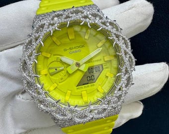 May include: A neon yellow G-Shock Casio watch with a diamond-encrusted bezel and band. The watch face displays the time and other functions. The watch is on a white surface.