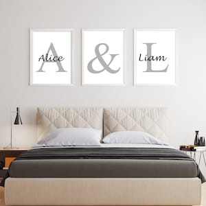May include: Three framed prints with the names "Alice" and "Liam" and an ampersand symbol. The names are in a script font, and the letters are in a gray color. The prints are above a bed with a beige headboard and a dark gray blanket.
