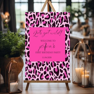 May include: A pink leopard print welcome sign on a wooden easel. The sign has a bright pink center with the text "Lets get wild!" and "Welcome to Ava's First Birthday Party 11.04.2026".
