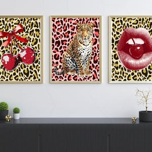 May include: Set of three framed art prints. One features red sequined cherries with a red bow on a leopard print background. Another shows a leopard on a pink leopard print. The third depicts red lips with a cherry on a leopard print background.