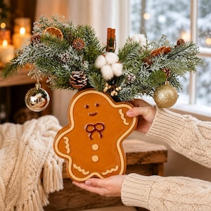 Può includere: Un vaso in ceramica a forma di omino di pan di zenzero riempito con una composizione festiva di rami di pino innevati, pigne, bastoncini di cannella, batuffoli di cotone e fette di arancia essiccata, con decorazioni natalizie dorate e argentate.
