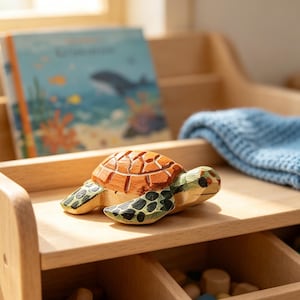 Könnte beinhalten: Handgeschnitzte Holzschildkrötenfigur mit orange-grünem Panzer, schwarz-grünen Flossen und detailliertem Design. Die Schildkröte liegt auf einem Holzregal, im Hintergrund ein Buch und eine blaue Decke.