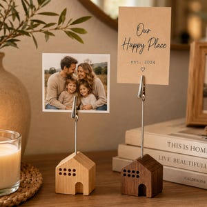 May include: Two house-shaped wooden photo holders with metal clips. One light wood and one dark wood base, each featuring a small arched doorway and window cutouts. The light holder displays a family photo, while the dark holder displays a tan card with the text Our Happy Place est. 2024 and a small heart symbol.