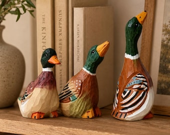 May include: Three hand-carved wooden duck figurines with visible chisel marks. The ducks feature painted details in shades of green, brown, orange, white, and black. Each duck is depicted in a different pose, with varying head angles and wing patterns.