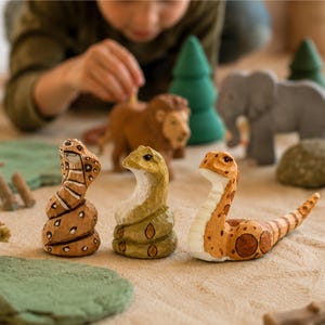 Peut inclure: Trois figurines de serpent en bois sculptées à la main alignées. Le serpent de gauche est brun clair avec des marques brun foncé. Celui du milieu est vert olive avec des taches vert foncé. Celui de droite est beige avec des taches brunes.