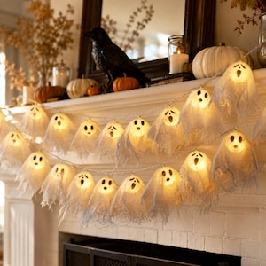 Puede incluir: Una guirnalda de luces LED con múltiples pequeñas fundas de tela blanca en forma de fantasma. Cada fantasma tiene una cara impresa con dos ojos oscuros y una boca abierta, iluminada desde el interior por una luz cálida y brillante.