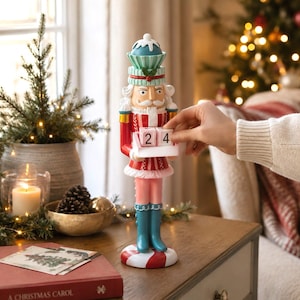 Può includere: Una statuetta schiaccianoci con un cappello color ottanio e bianco, una giacca rossa con bordi bianchi e pantaloni rosa. La figura regge un vassoio bianco con due blocchi di legno che mostrano il numero 24, su una base a righe rosse e bianche.