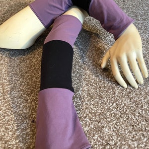 May include: A pair of arm warmers made of stretchy fabric. The arm warmers are purple, black, and white. They are shown on a mannequin arm.