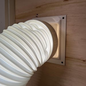 May include: A white, flexible, ribbed ventilation hose is connected to a white square vent. The vent is mounted on a light brown wooden surface. The hose curves to the left, showing its accordion-like structure.