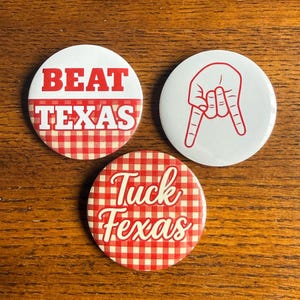 May include: Three round buttons with red and white designs. One button says "BEAT TEXAS" in red letters over a red and white checkered pattern. Another button has a red hand gesture. The third button says "Tuck Texas" in red letters over a red and white checkered pattern.