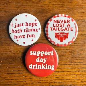 May include: Three round buttons with text. One white button says "i just hope both teams have fun" with red stars. Another button says "NEVER LOST A TAILGATE" with red cups on a white background with a red and white checkered border. The third button says "support day drinking".