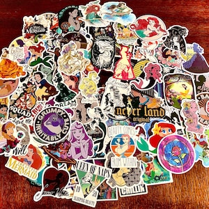 Könnte beinhalten: Eine Sammlung bunter Aufkleber mit verschiedenen Disney-Charakteren und Zitaten. Die Aufkleber zeigen Charaktere aus Die Schöne und das Biest, Die kleine Meerjungfrau, Schneewittchen und mehr. Die Aufkleber sind auf einer Holzoberfläche angeordnet.