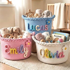 Puede incluir: Tres cestas de almacenamiento redondas de cuerda tejida en rosa, azul y blanco. Cada cesta presenta un borde trenzado, un detalle de flor de ganchillo y un nombre personalizado en letras de lana de colores: Emily, Lucas y Olivia. Las cestas contienen varios juguetes para bebés, peluches y libros de cartón.
