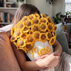 Op de afbeelding: Een gehaakt boeket met talloze gele en bruine zonnebloemen bevestigd op een witte basis. Het witte gedeelte heeft de naam Mae in geel garen op de voorkant geborduurd. De bloemen zijn dicht bezet met bloemblaadjes met een textuur.