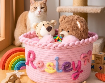 Könnte beinhalten: Ein rosa Seilkorb mit dem Namen "Resley" in bunten Garnbuchstaben. Der Korb enthält Stofftiere, darunter ein Teddybär und ein Leopard. Ein Holzregenbogen, ein Spielzeugauto und Blöcke befinden sich in der Nähe.