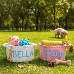 Puede incluir: Dos cestas de cuerda sobre hierba, una blanca con "BELLA" en letras azules, con un juguete de cerdo rosa y una pelota azul. La otra es rosa con "Nancy" en azul, con una toalla amarilla y un juguete de cuerda. Un perro juega al fondo.