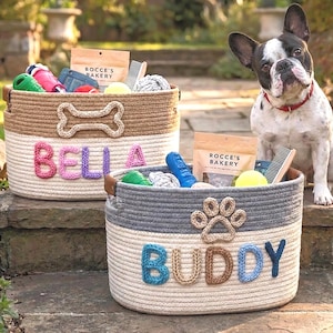 Puede incluir: Dos cestas de almacenamiento redondas de cuerda trenzada llenas de artículos para mascotas. La cesta beige y blanca presenta una forma de hueso y el nombre BELLA en rosa y morado. La cesta gris y blanca presenta una huella y el nombre BUDDY en azul y beige.