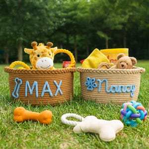 Puede incluir: Dos cestas de cuerda con letras azules "MAX" y "Nancy" sobre hierba verde. La cesta "MAX" contiene un peluche de jirafa. La cesta "Nancy" contiene un peluche de oso de peluche. Juguetes para perros esparcidos delante.