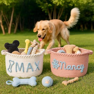 Puede incluir: Dos cestas de cuerda llenas de juguetes para perros sobre hierba. Una es de color crema con "MAX" en letras azules, la otra es rosa con "Nancy" en escritura azul. Un perro golden retriever est&aacute; en el fondo.