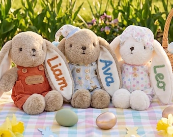 May include: Three plush bunny rabbits with personalized names embroidered on their ears. One bunny wears orange overalls, another a blue pajama set, and the third a white polka-dot nightgown. Easter eggs and a basket are in the background.