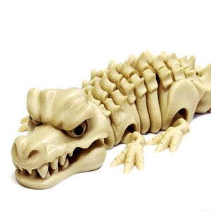 May include: A beige, articulated dragon figurine with a detailed, textured design. The dragon has a menacing expression, sharp teeth, and a spiky spine. The figurine is lying down on a white surface.