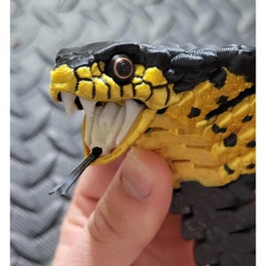Könnte beinhalten: Ein detaillierter, 3D-gedruckter Schlangenkopf mit einem schwarz-gelben Farbschema. Das Maul der Schlange ist geöffnet und zeigt weiße Zähne und eine schwarze gespaltene Zunge. Das Auge ist hellbraun. Der Schlangenkopf wird von einer Hand gehalten.