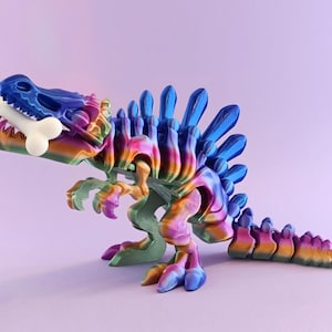 Op de afbeelding: Een kleurrijk 3D-geprint dinosaurusbeeldje met een bot in zijn bek. De dinosaurus heeft een regenboogkleurverloop, waaronder blauw, groen, geel en roze. Het beeldje staat op een lichtpaarse ondergrond.