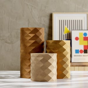 May include: Three geometric paper vases in varying sizes and shades of brown. The vases have a faceted design, creating a textured appearance. The background includes framed art and a neutral wall.