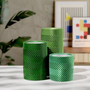 May include: Three green cylindrical planters with a textured chevron pattern. The planters are different sizes, with the largest in the center and the smallest in the front. The planters are on a white surface, with a plant and artwork in the background.