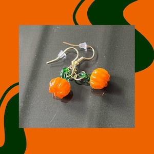 以下が含まれることがあります： 小さなオレンジ色のカボチャのチャームが付いた、金色のイヤリングのペア。カボチャは質感のある表面で、グリーンのガラスアクセントでイヤリングフックに接続されています。イヤリングは暗い背景に配置されています。