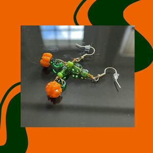 以下が含まれることがあります： オレンジ色のカボチャのチャームと緑と透明なガラスビーズが特徴の手作りのイヤリング。 イヤリングは銀色のイヤーフックが付いており、秋の季節にぴったりのアクセサリーです。