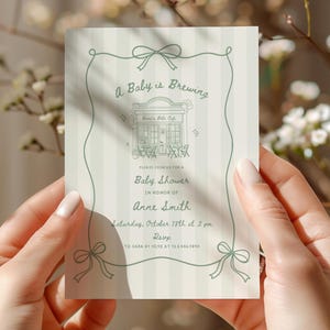 May include: A rectangular baby shower invitation card with a light green striped background and a sage green border featuring bows. The text reads A Baby is Brewing, Anne's Tea Cafe, Baby Shower in honour of Anne Smith, Saturday, October 18th at 2 pm.