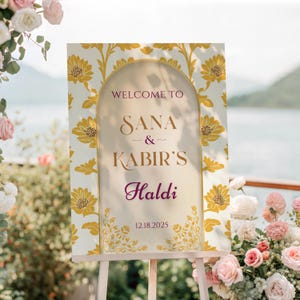 May include: A wedding welcome sign on an easel. The sign has a cream background with a yellow floral pattern. The text reads "Welcome to Sana & Kabir's Haldi" and the date 12.18.2025.